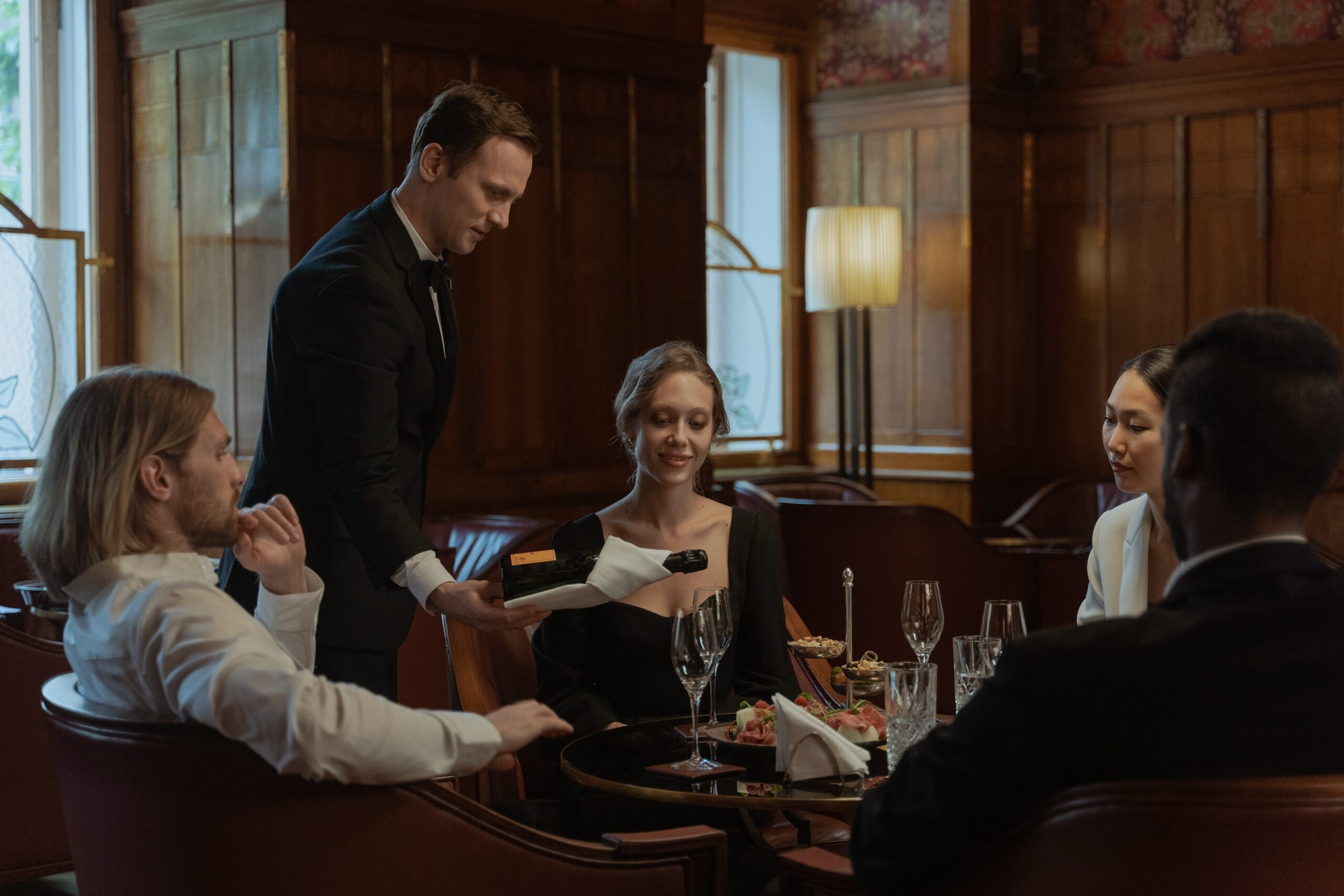 A waiter pours a wine from the bottle to a girl, sitting with her friends in the restaurant.