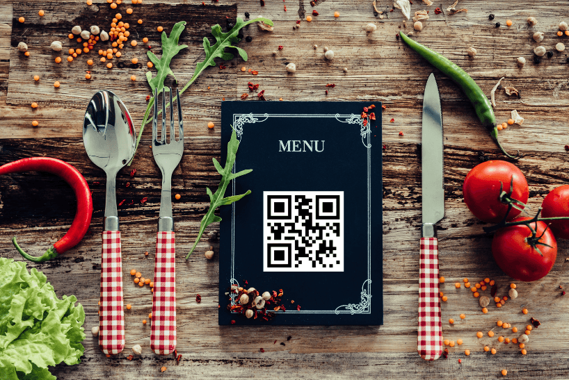 A menu with a QR code on it is placed on the table with kitchen utensils and some vegetables.