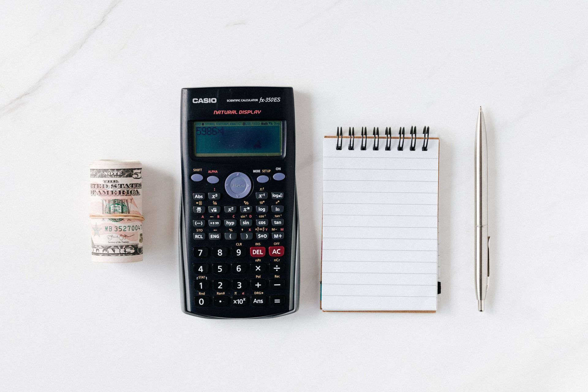 A dollar, calculator, and notebook with a pen are placed next to each other.
